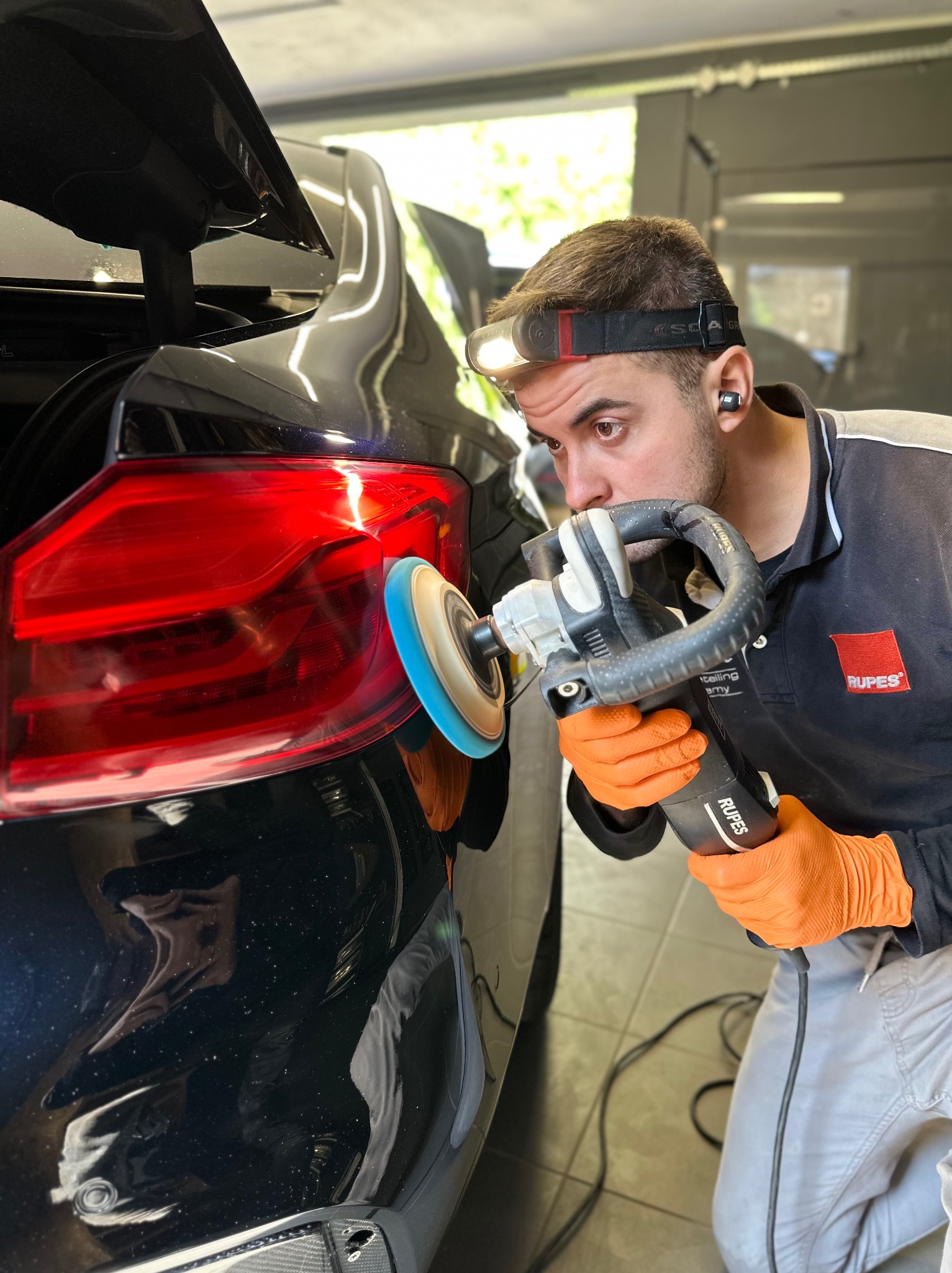 Detailer w pomarańczowych rękawicach poleruje tylną lampę czarnego samochodu przy użyciu polerki Rupes, oświetlając sobie pracę lampą czołową.