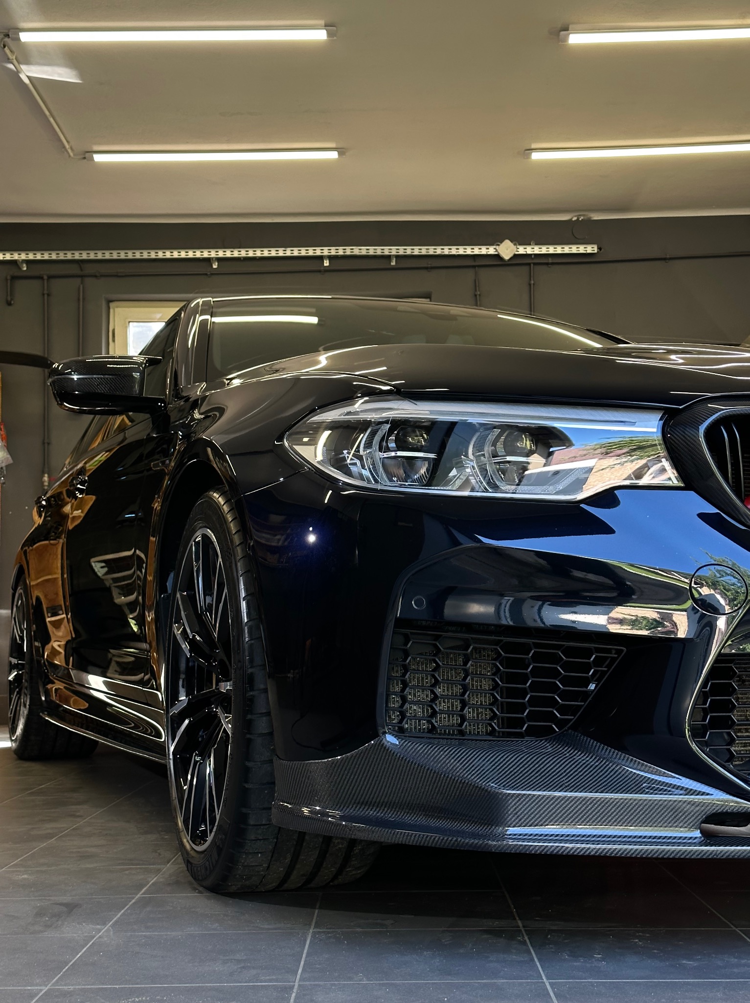 Błyszczący, czarny samochód sportowy z karbonowymi elementami, sfotografowany pod niskim kątem w dobrze oświetlonym garażu z szarą podłogą i ścianami.