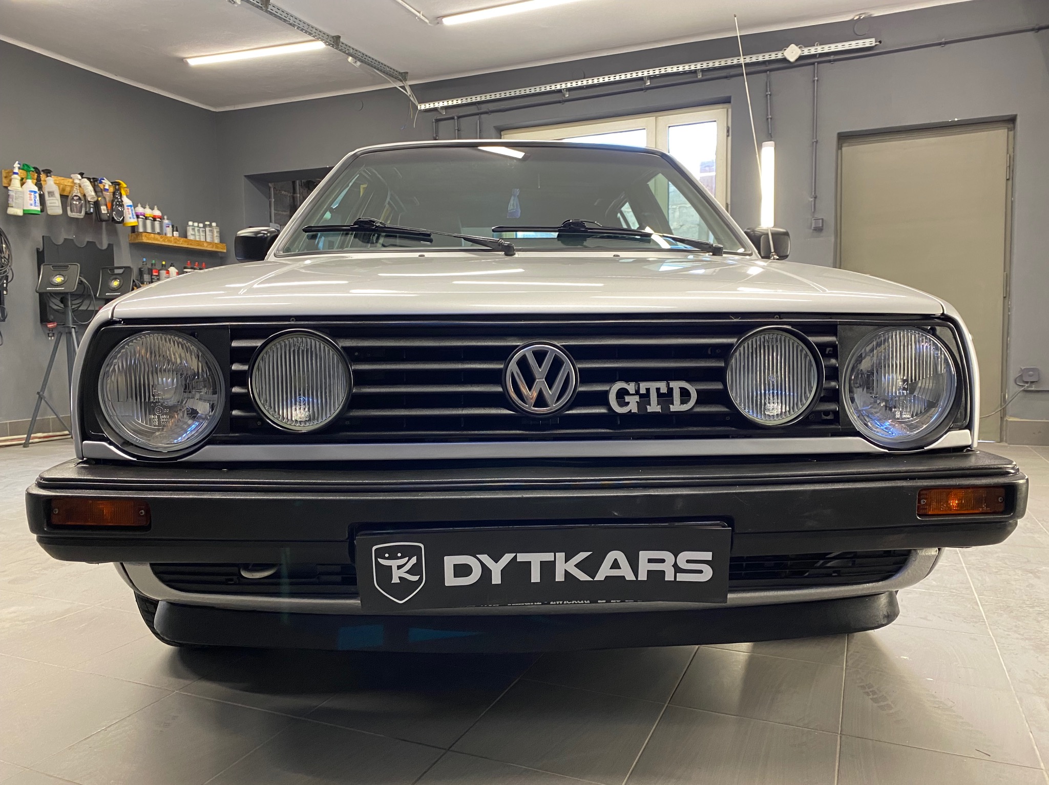Przód białego samochodu VW GTD z logo firmy DYTKARS na tle garażu z półkami pełnymi środków czyszczących.