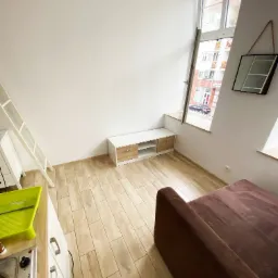 Wnętrze jasnego pokoju z wysokim oknem, widoczne białe ściany, drewnopodobne panele, sofa w kolorze ciemnego brązu, biała drabina, szafka RTV i akcesoria malarskie.