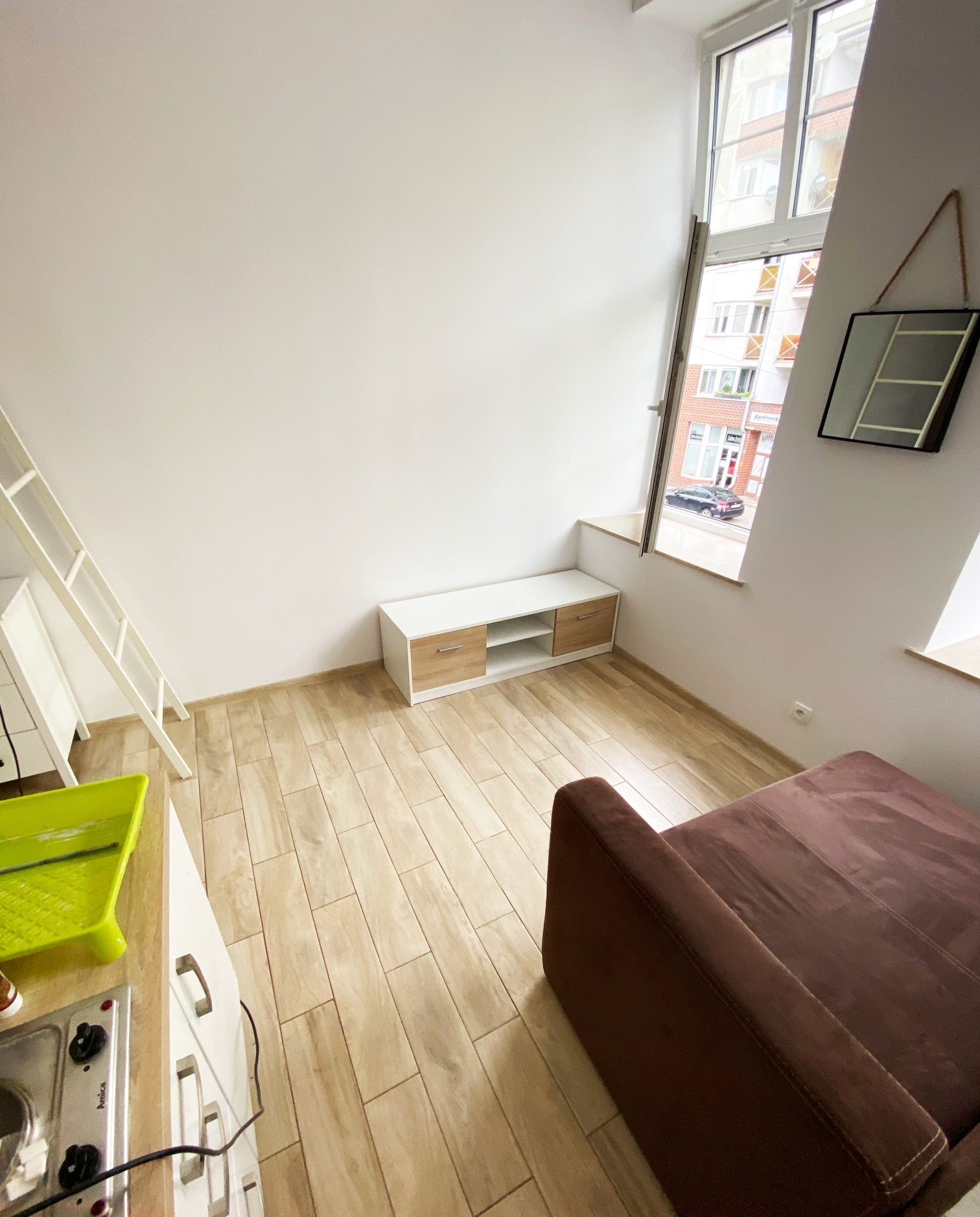 Wnętrze jasnego pokoju z wysokim oknem, widoczne białe ściany, drewnopodobne panele, sofa w kolorze ciemnego brązu, biała drabina, szafka RTV i akcesoria malarskie.