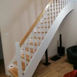 Drewniane schody z białymi balustradami i oświetleniem LED w stopniach, widok z dołu na zakrzywiony fragment balustrady.