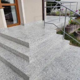 Schody wejściowe do domu pokryte jasną, nakrapianą posadzką żywiczną z metalową barierką, otoczone zielenią i kwiatami w betonowych donicach.