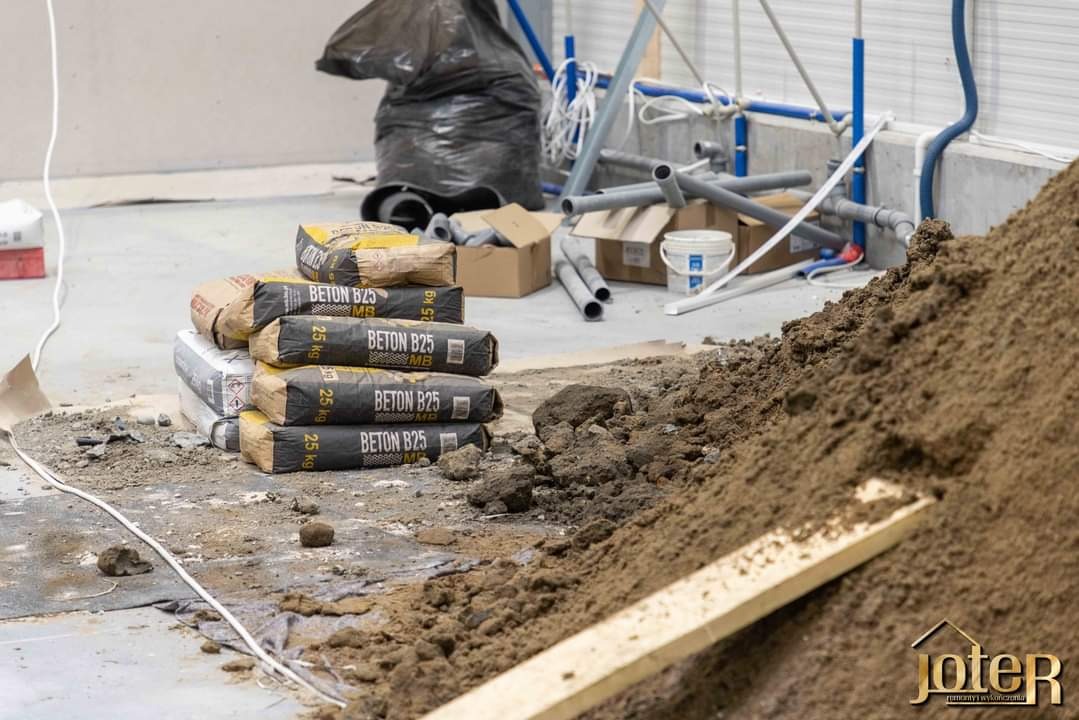 Stos worków z betonem B25 na budowie, obok hałda ziemi, rury PCV i inne materiały budowlane, widoczne logo firmy Joter w prawym dolnym rogu.