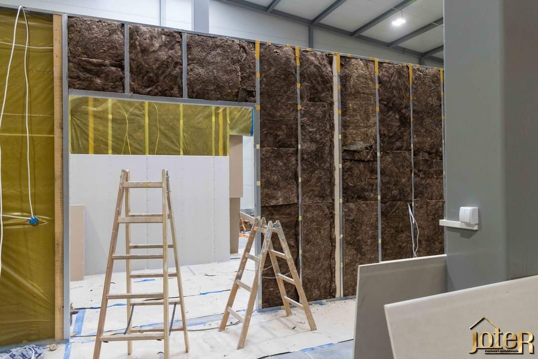 Montaż izolacji z wełny mineralnej w metalowej konstrukcji ścian działowych, widoczne drewniane drabiny i folia paroizolacyjna, w tle białe płyty gipsowo-kartonowe.