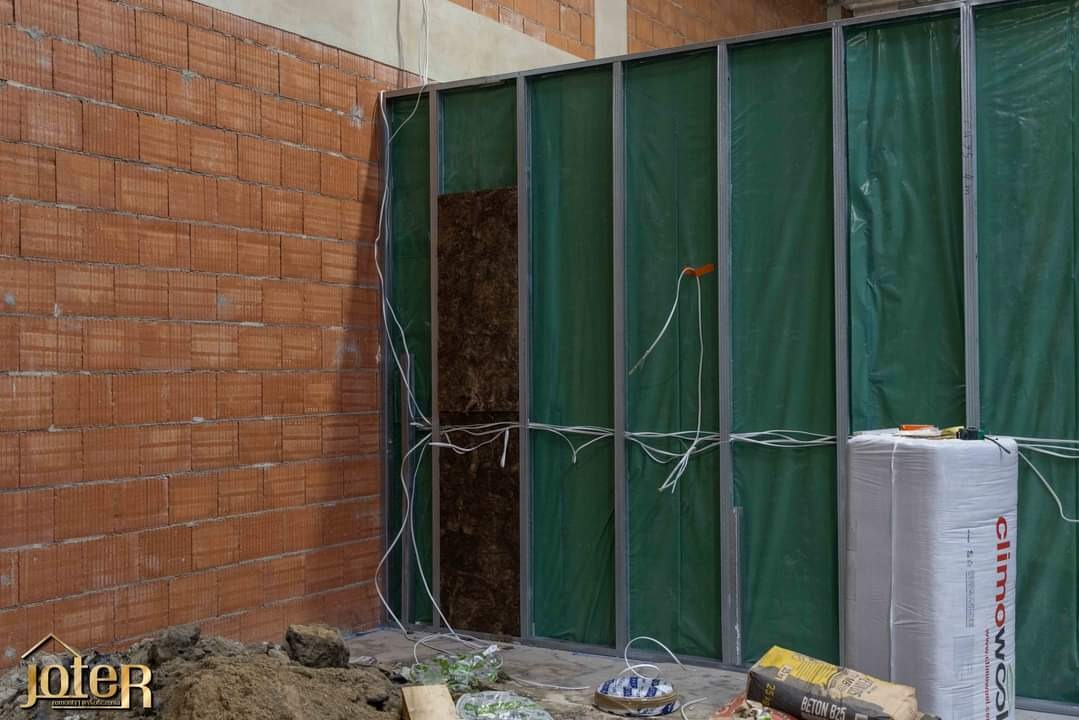 Wnętrze w trakcie prac: widoczna ściana z czerwonej cegły, metalowa konstrukcja z zieloną folią i częściowo zamontowaną izolacją, kable elektryczne oraz worki z zaprawą murarską na podłodze.