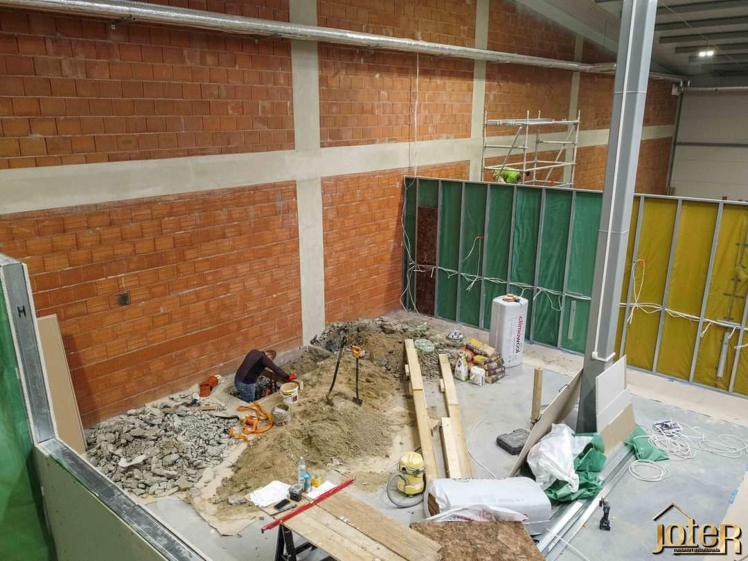 Wnętrze w trakcie prac budowlanych, widoczne ceglane ściany, metalowe konstrukcje szkieletowe z zielonymi i żółtymi panelami, gruz, piasek, narzędzia i pracownik w trakcie wykonywania prac ziemnych...