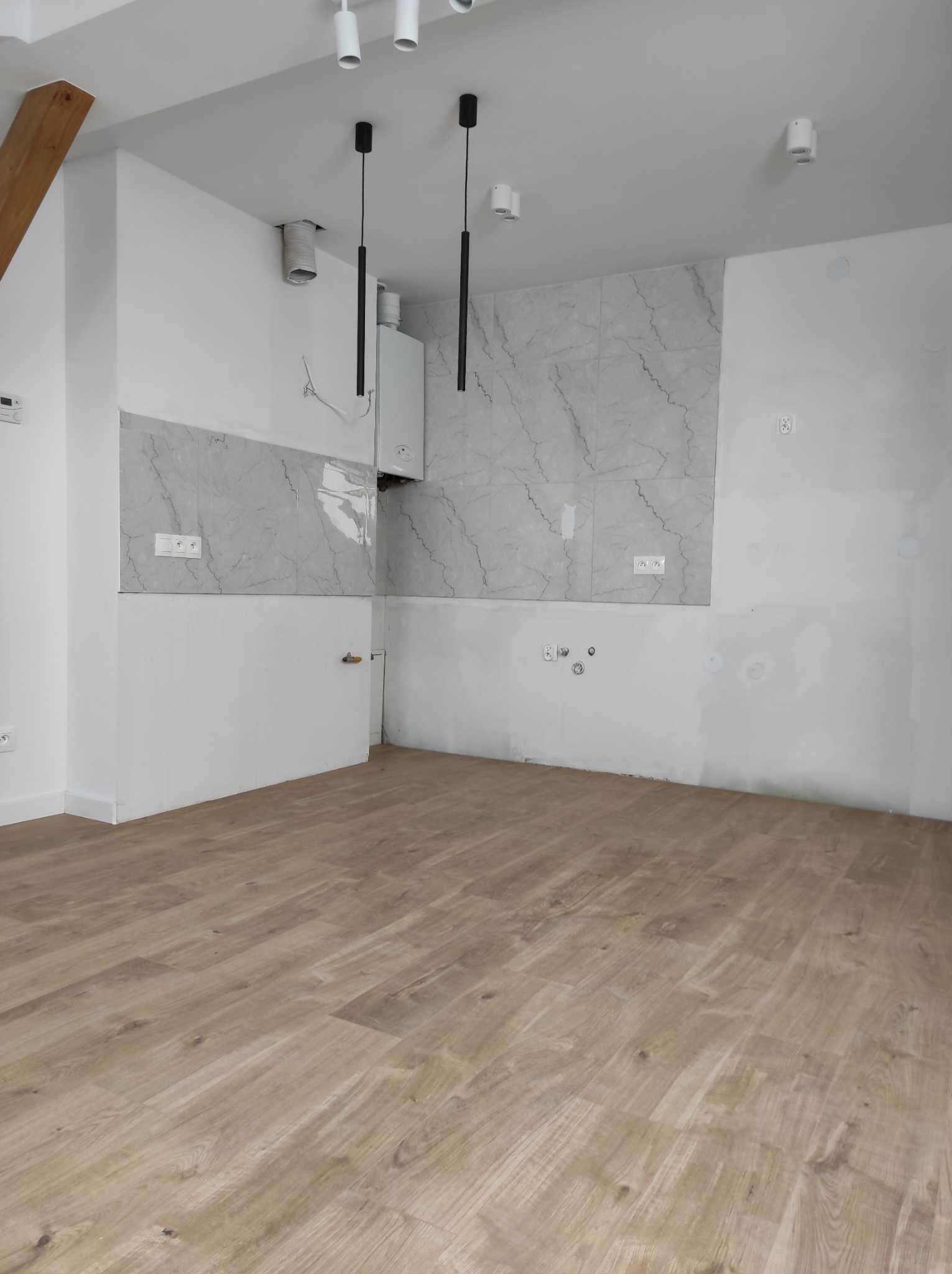 Wnętrze w trakcie prac wykończeniowych: panele podłogowe ułożone, ściany częściowo z płytkami imitującymi marmur, widoczny piec gazowy i instalacja elektryczna.