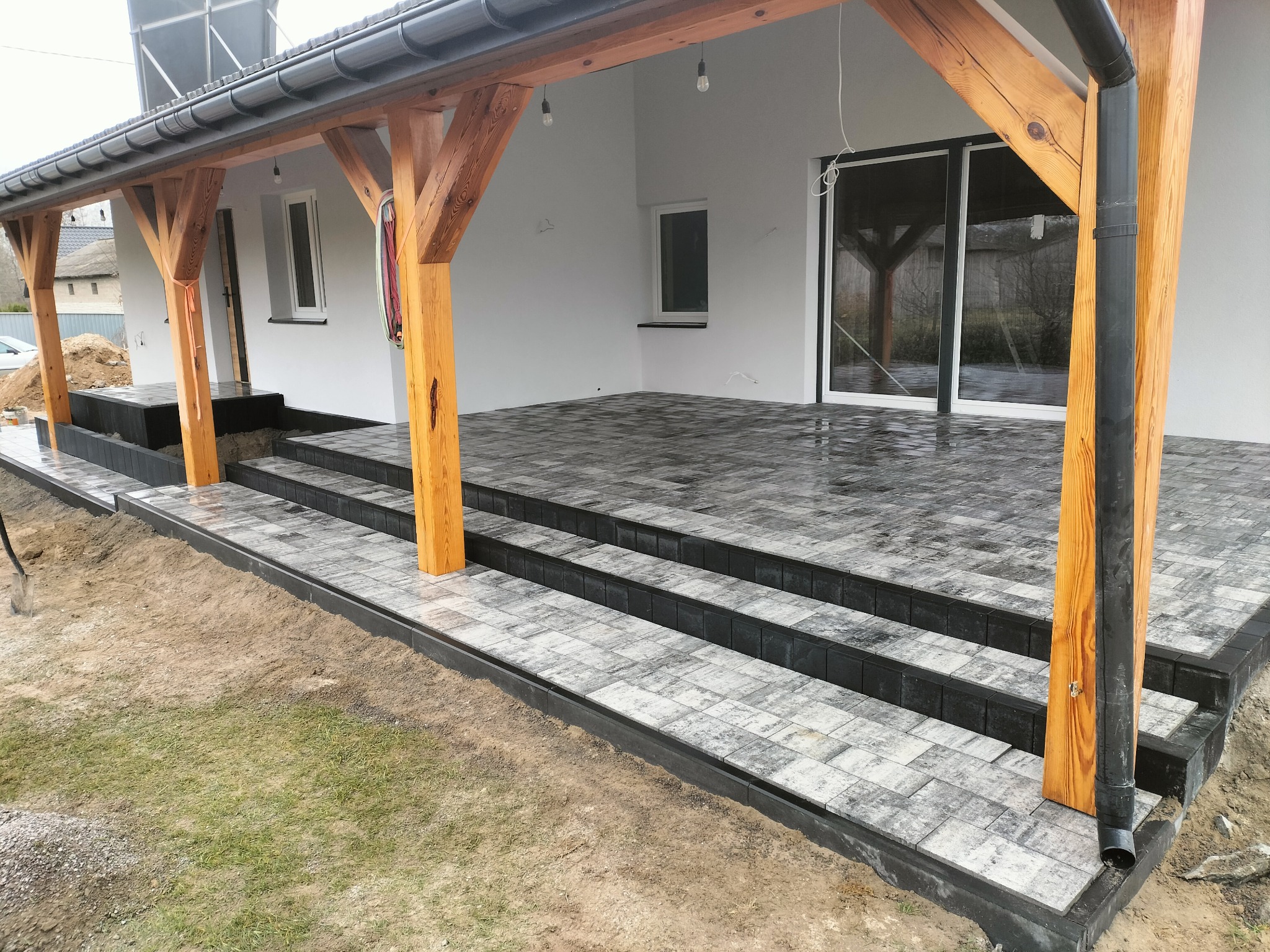 Zadaszony taras z kostki brukowej w odcieniach szarości, z drewnianymi słupami i czarnymi rynnami. Widoczne trzy stopnie i fragment trawnika.