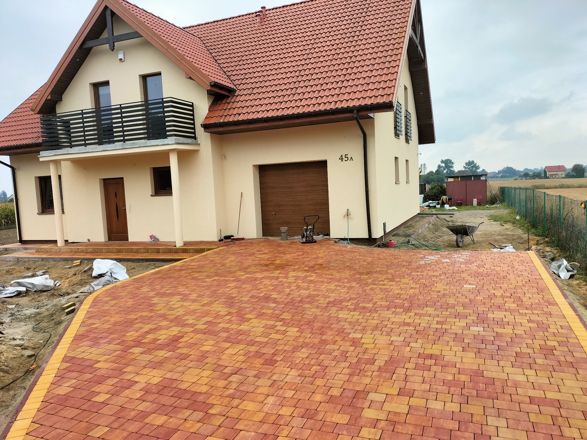Nowa, pomarańczowo-żółta kostka brukowa przed domem w Siedliszczach. Widoczne narzędzia brukarskie i fragment domu z balkonem, co sugeruje świeżo zakończone prace.