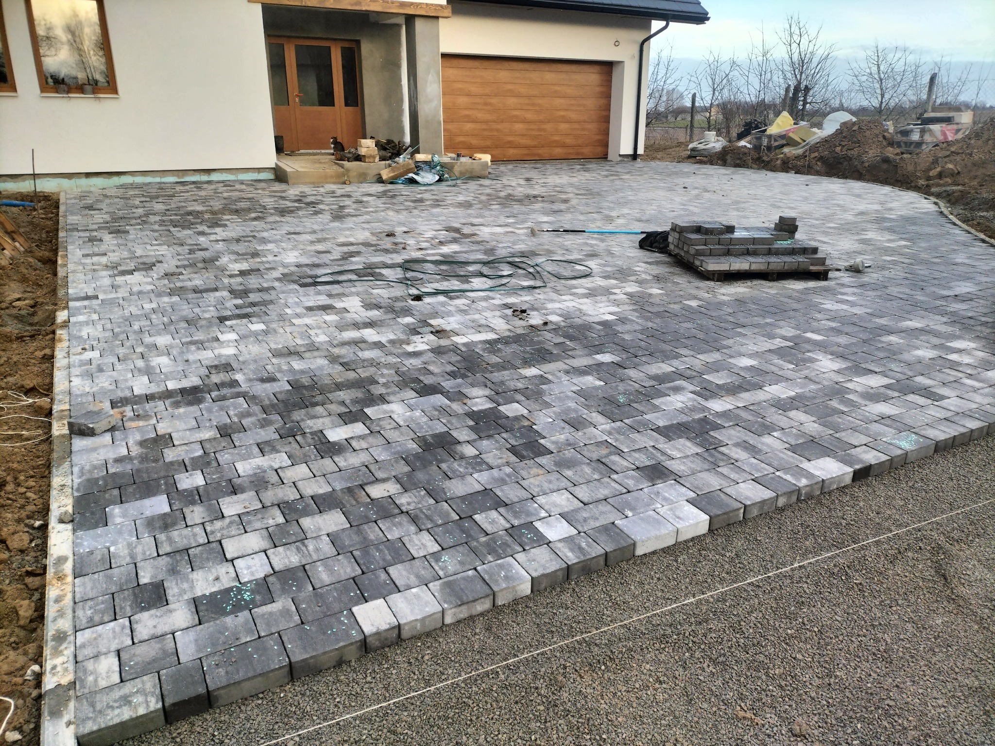 Ułożona, szara kostka brukowa na podjeździe domu jednorodzinnego. Widoczne niedokończone krawędzie i przygotowany teren pod dalsze prace brukarskie.