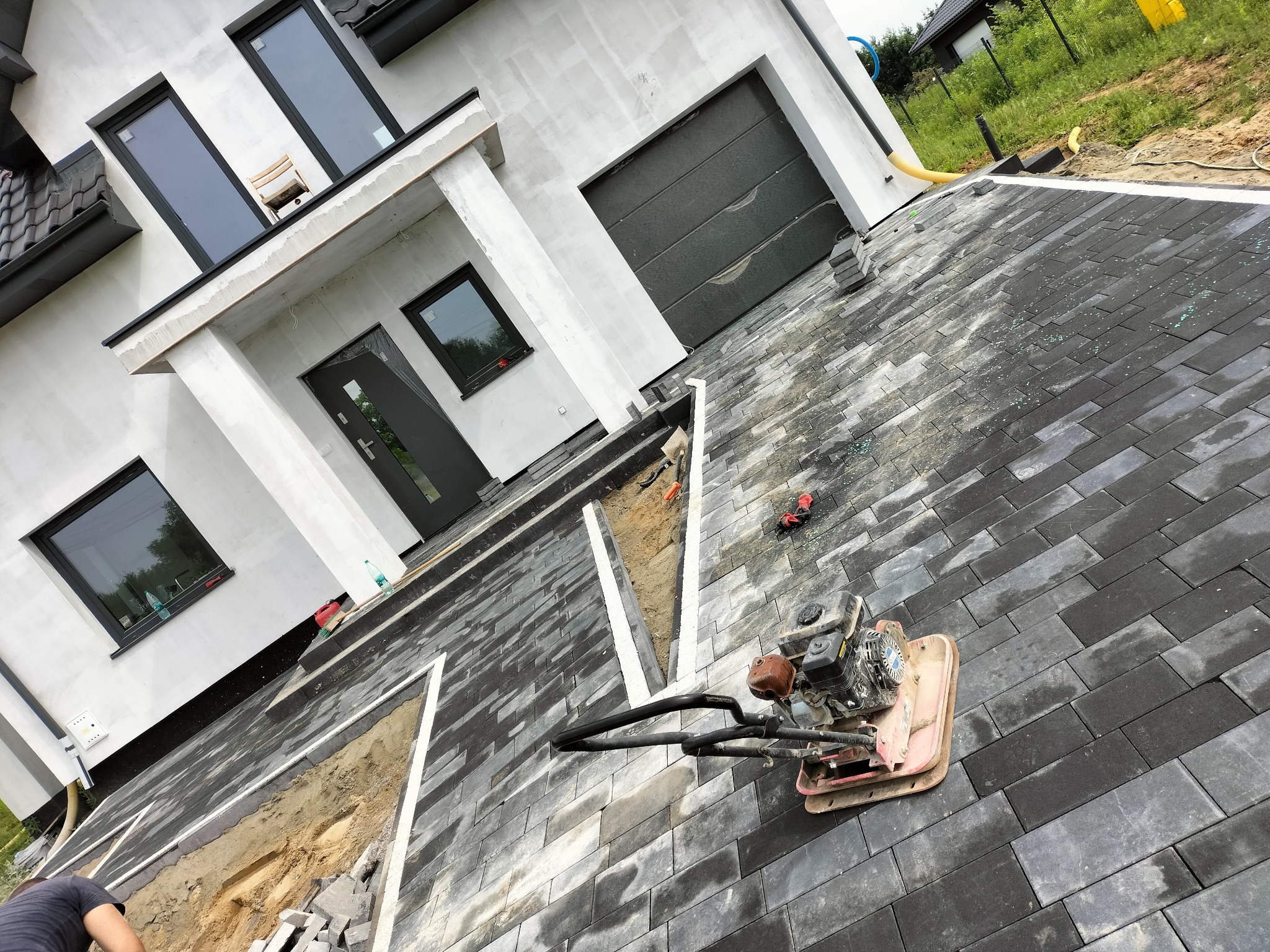 Układanie szarej kostki brukowej prostokątnej na posesji. Wibrator do ubijania kostki. Fragment budynku z szarą stolarką okienną i drzwiową.