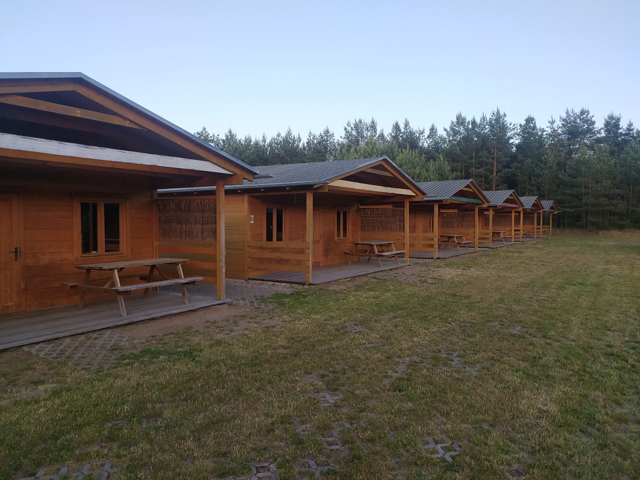 Szereg identycznych, drewnianych domków letniskowych z zadaszonymi tarasami i drewnianymi stolikami piknikowymi, ustawionych w rzędzie na trawiastym terenie w otoczeniu lasu iglastego.