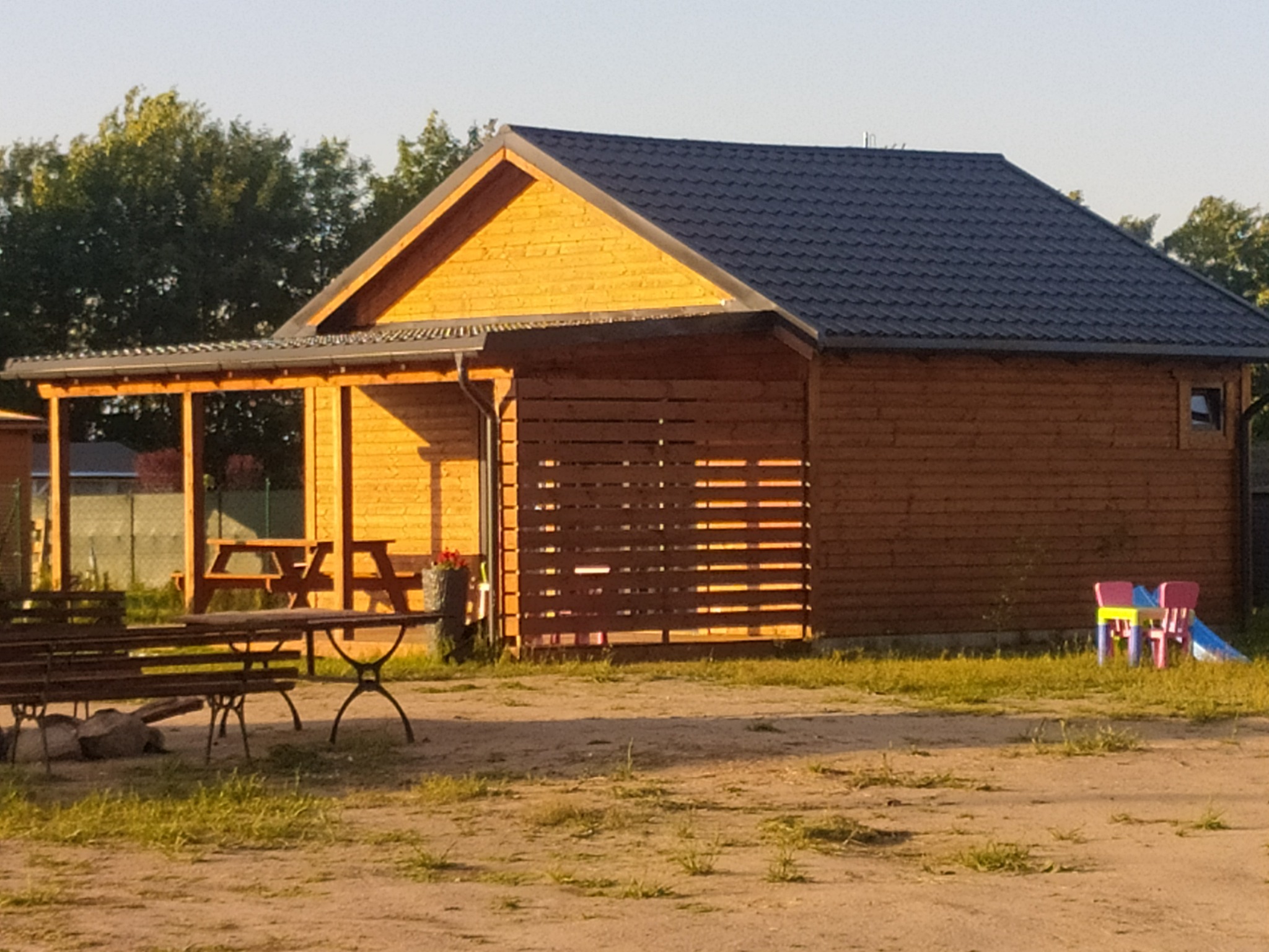 Drewniana konstrukcja z zadaszeniem i ażurową ścianą, prawdopodobnie domek letniskowy lub altana, z ciemną dachówką, w tle drzewa i dziecięce krzesełka.