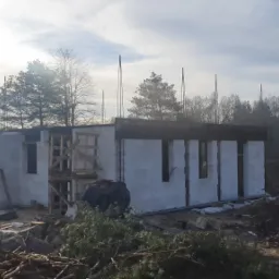 Budowa domu murowanego w stanie surowym otwartym, widoczne białe bloczki, szalunki okienne, wystające pręty zbrojeniowe, ziemia i gałęzie w pierwszym planie, drzewa w tle, słoneczny dzień.