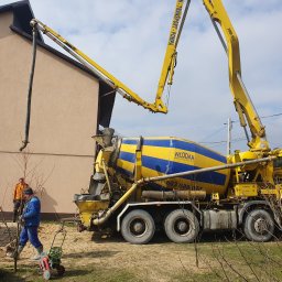 LUX-MIX ARTUR KIJAK - Żółto-niebieska betoniarka z wysięgnikiem pompuje beton do fundamentów domu jednorodzinnego, widoczni dwaj pracownicy budowlani oraz zbrojenie fundamentu.