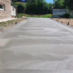 Świeżo wylana, gładka powierzchnia betonowa na podjeździe, z widocznymi śladami poziomowania, zabezpieczona pomarańczową taśmą ostrzegawczą, otoczona ziemią i elementami drewnianymi.