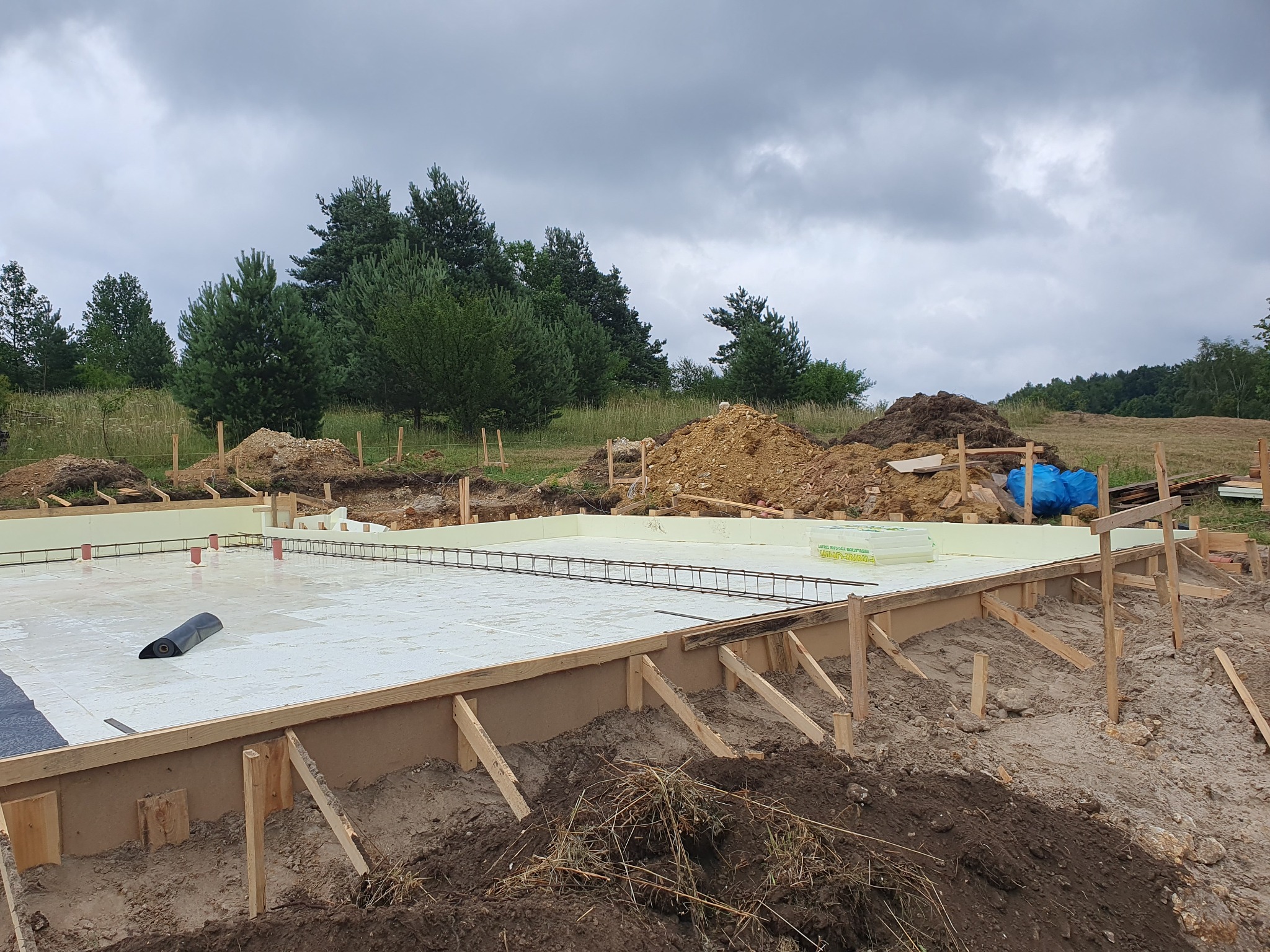Przygotowany szalunek z izolacją termiczną i zbrojeniem pod płytę fundamentową na działce z drzewami w tle.