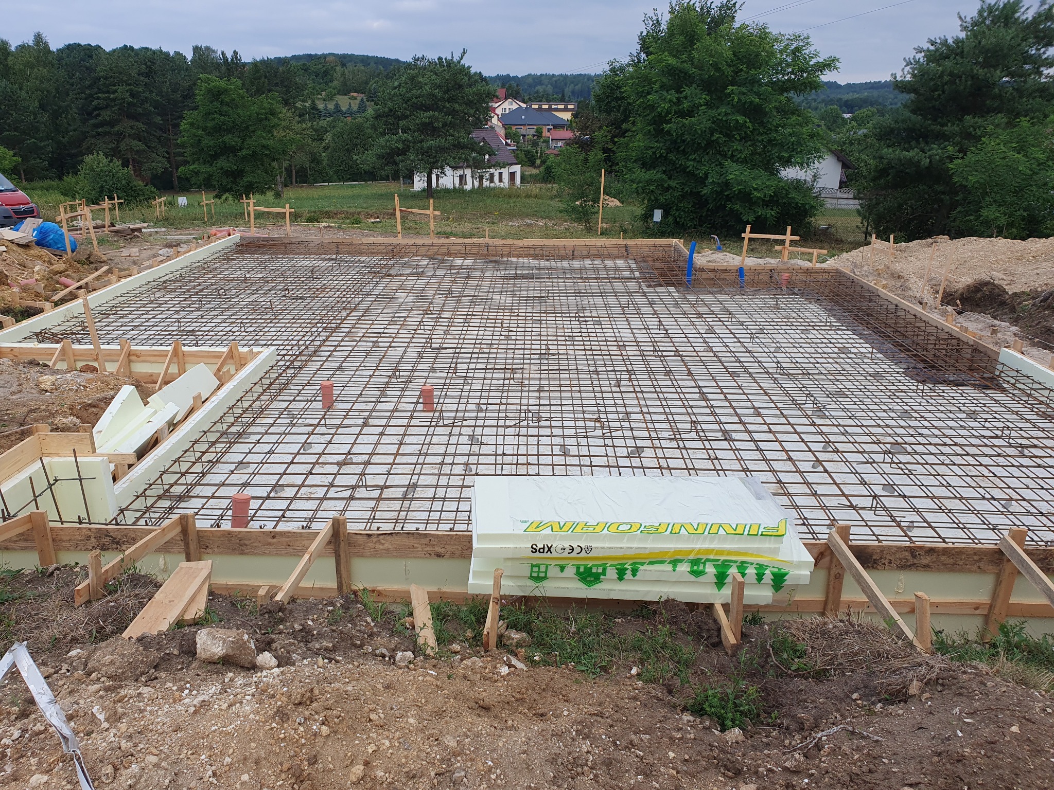 Zbrojenie fundamentu domu jednorodzinnego z widoczną siatką zbrojeniową, szalunkiem z drewnianych desek i styropianu, przygotowane do wylania betonu na tle zieleni.