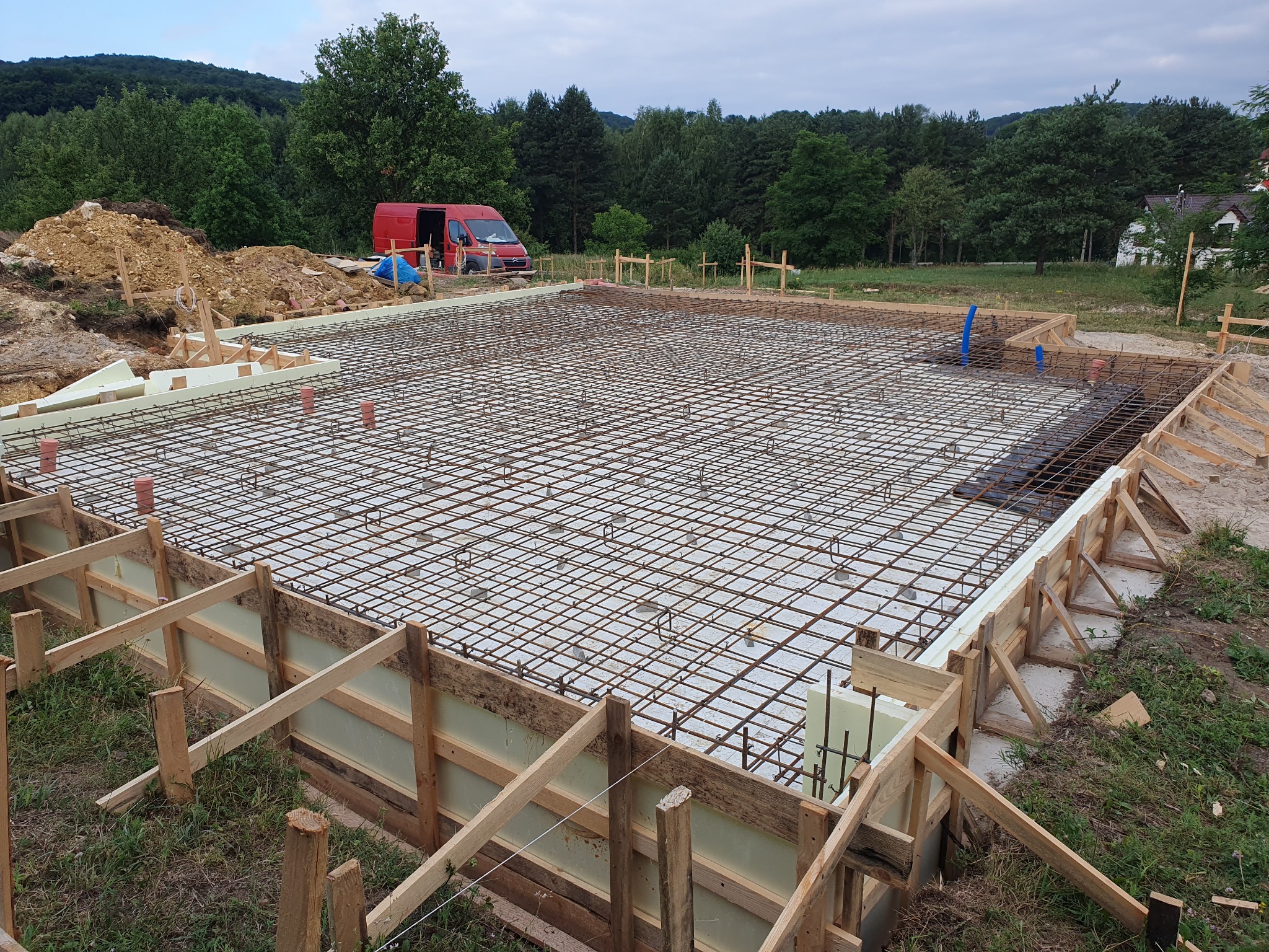 Przygotowany szalunek z wypełnieniem styropianowym i ułożone zbrojenie stalowe na płycie fundamentowej domu jednorodzinnego, widoczny czerwony samochód dostawczy w tle.