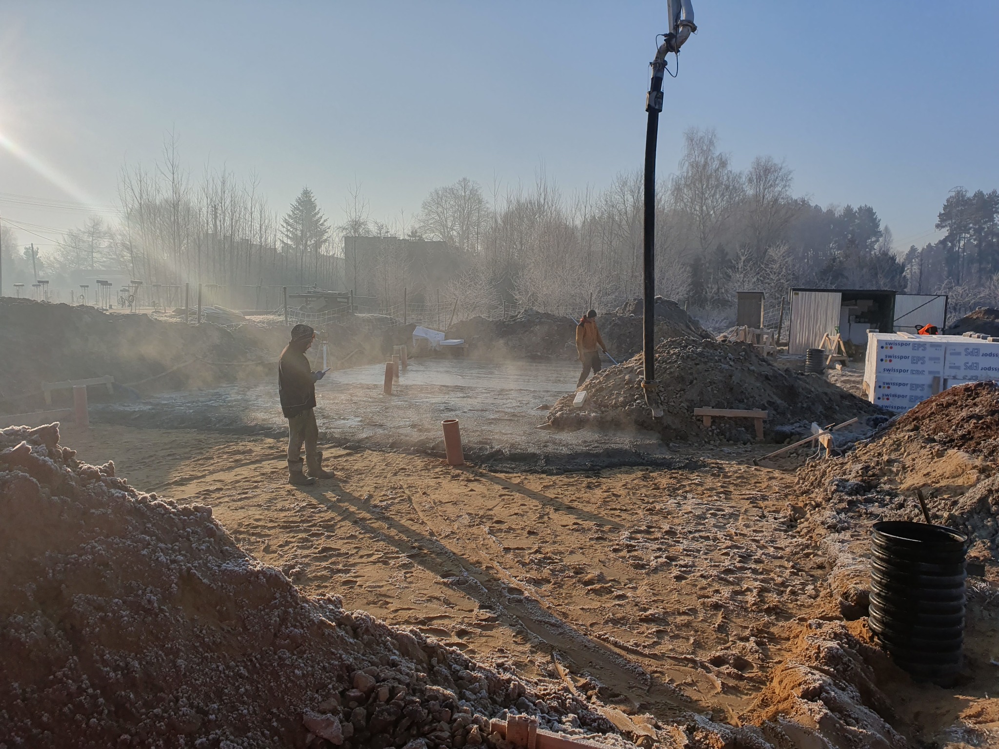 Przygotowanie terenu pod fundamenty w zimowy poranek, widoczna pompa do betonu, pracownicy oraz zapasy materiałów budowlanych.