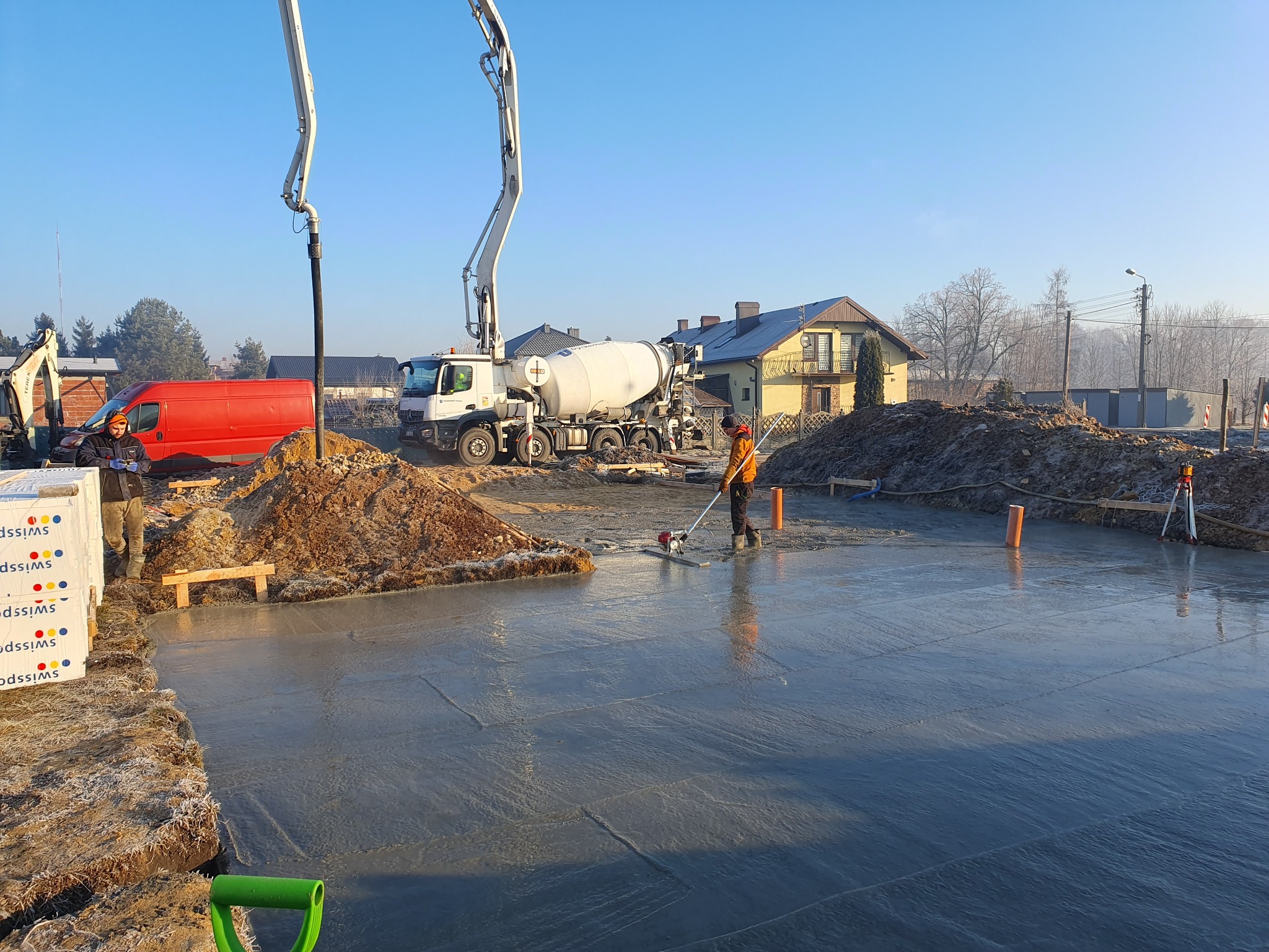 Wylewanie betonu na przygotowany teren pod budowę domu jednorodzinnego; widoczny mikser do betonu, w tle dom w trakcie budowy, pracownik zacierający powierzchnię betonu.