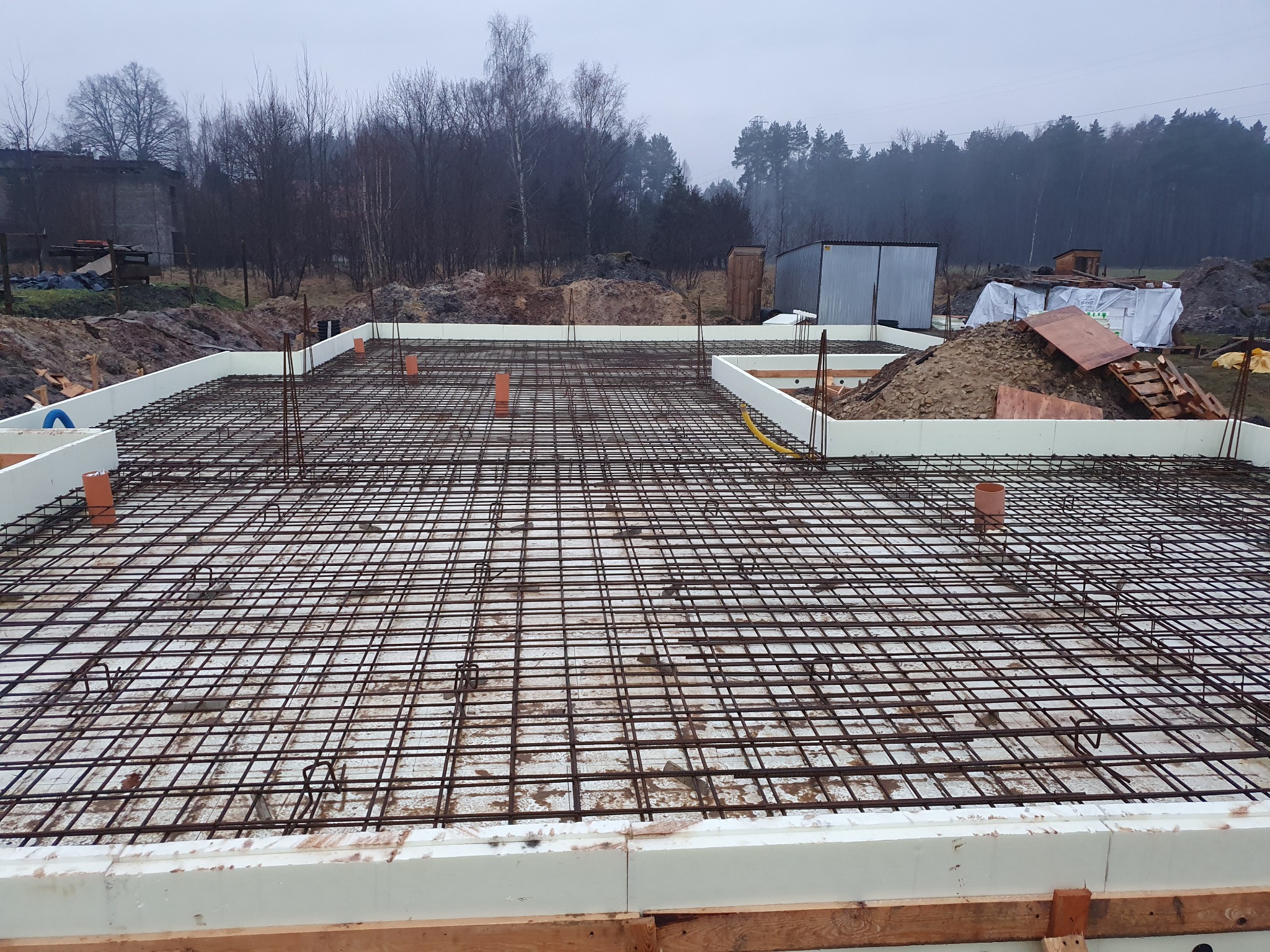 Siatka zbrojeniowa ułożona na styropianie w szalunku fundamentowym, widoczne rury kanalizacyjne, w tle drzewa i budynek gospodarczy.