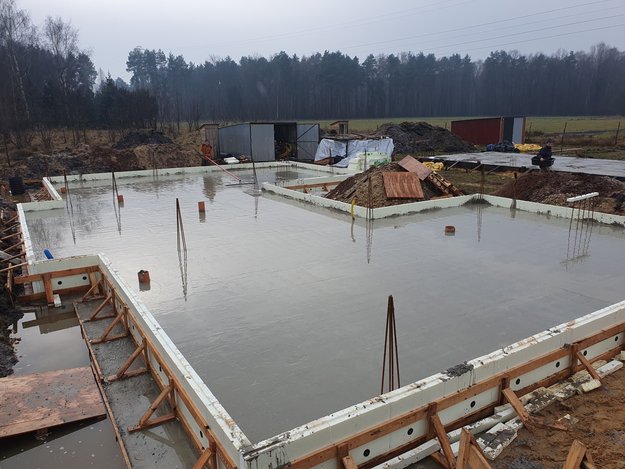 Świeżo wylany beton na płycie fundamentowej w szalunkach z białego styropianu, widoczne wystające pręty zbrojeniowe, w tle budynki gospodarcze i las w pochmurny dzień.