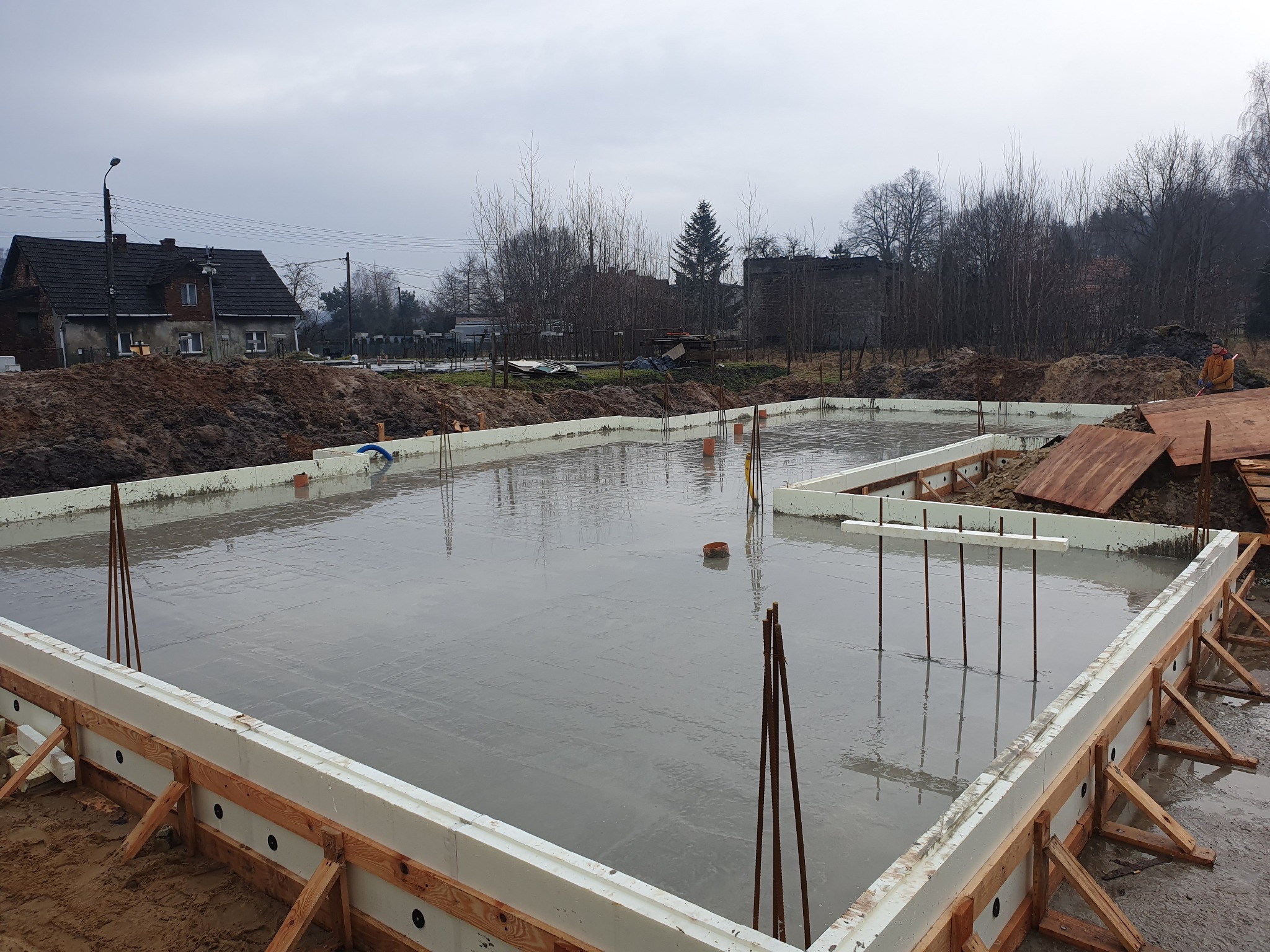 Wylany fundament z szalunkiem styropianowym i wystającymi prętami zbrojeniowymi, widok z góry na plac budowy w pochmurny dzień.