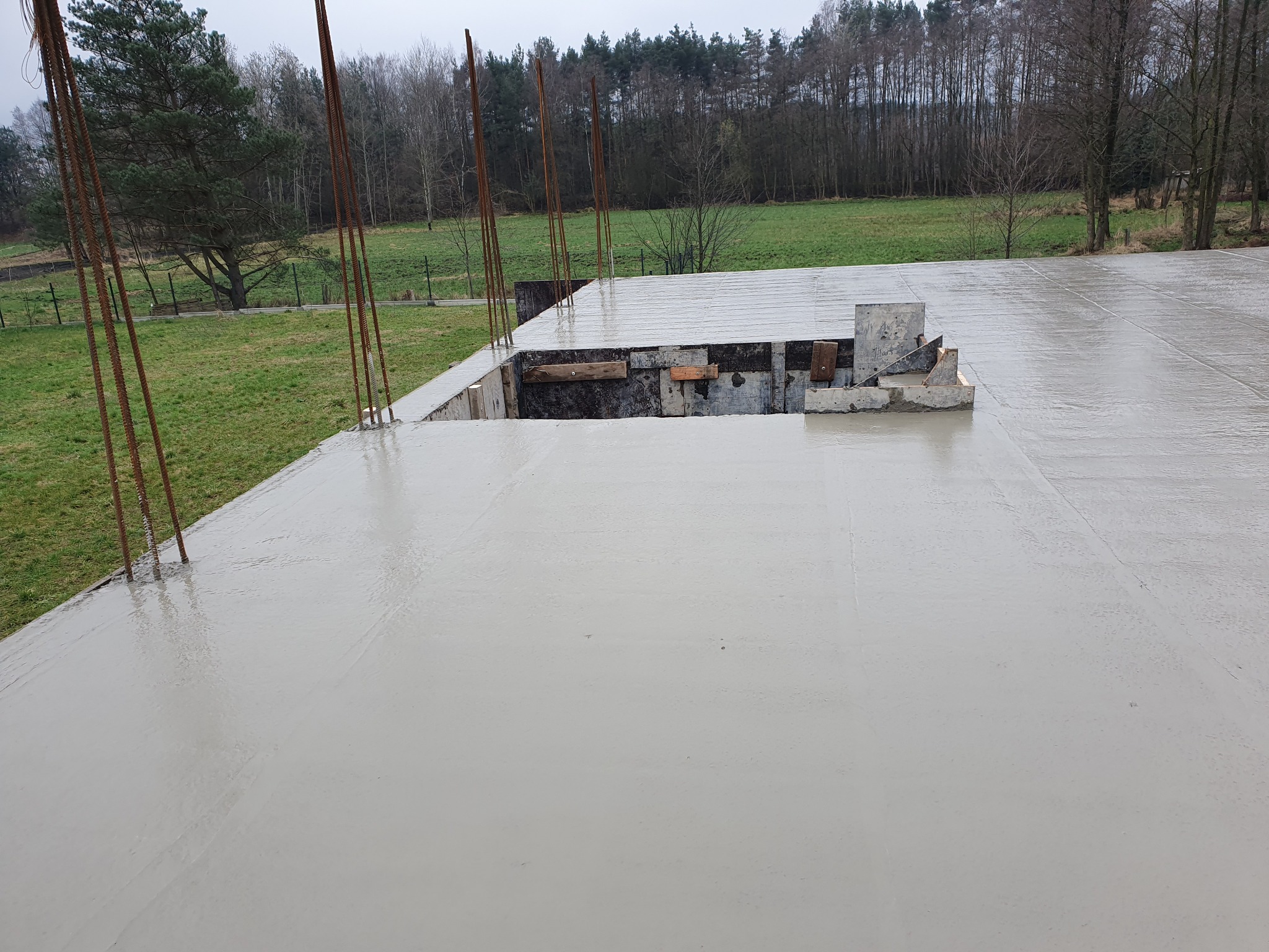 Świeżo wylana, gładka płyta fundamentowa z wystającymi prętami zbrojeniowymi, otoczona drewnianym szalunkiem, na tle zielonego krajobrazu z drzewami.