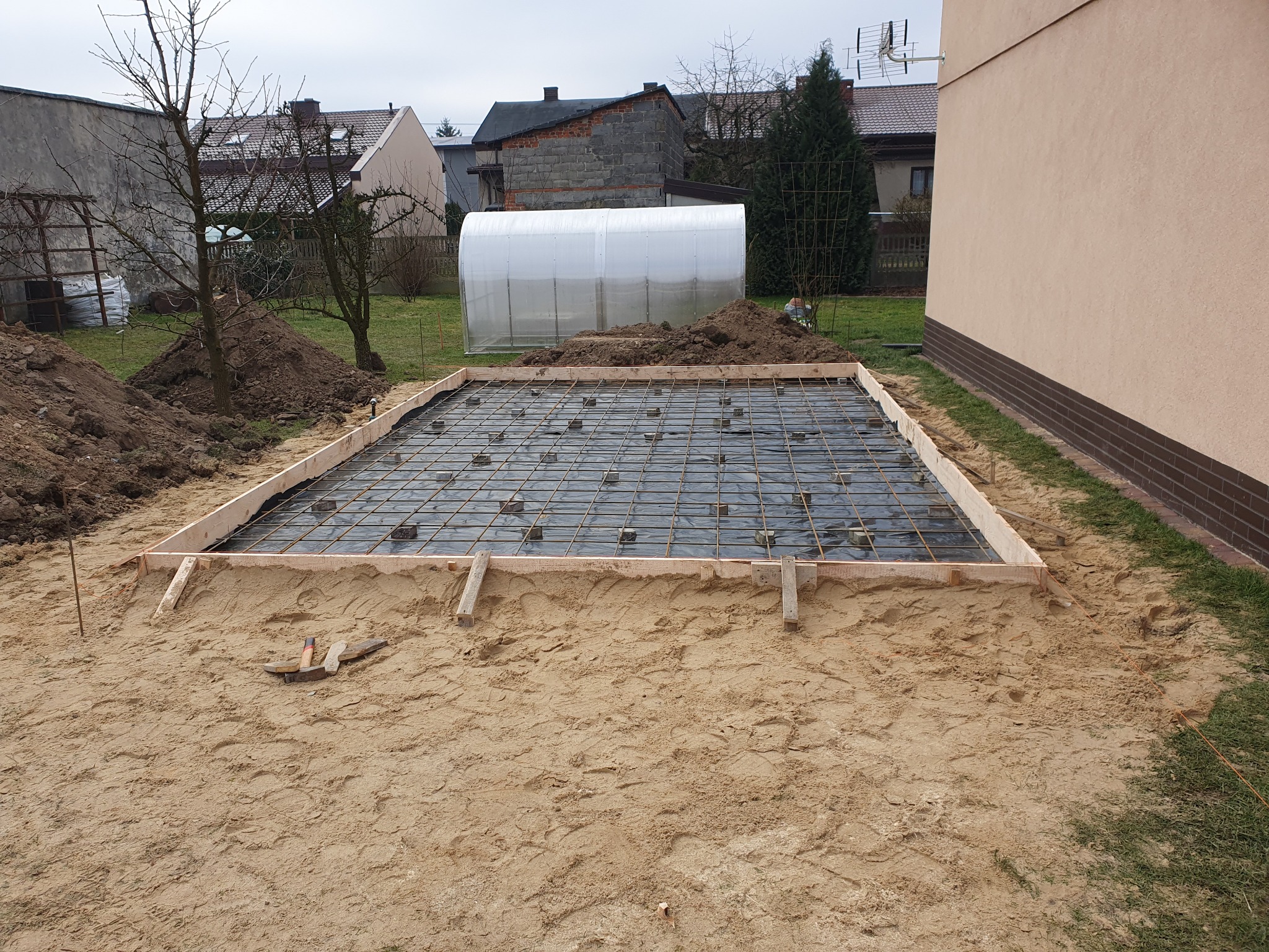 Przygotowany szalunek z siatką zbrojeniową i folią pod wylewkę fundamentową na piaszczystym podłożu, w tle ogród z tunelem foliowym i zabudowania.