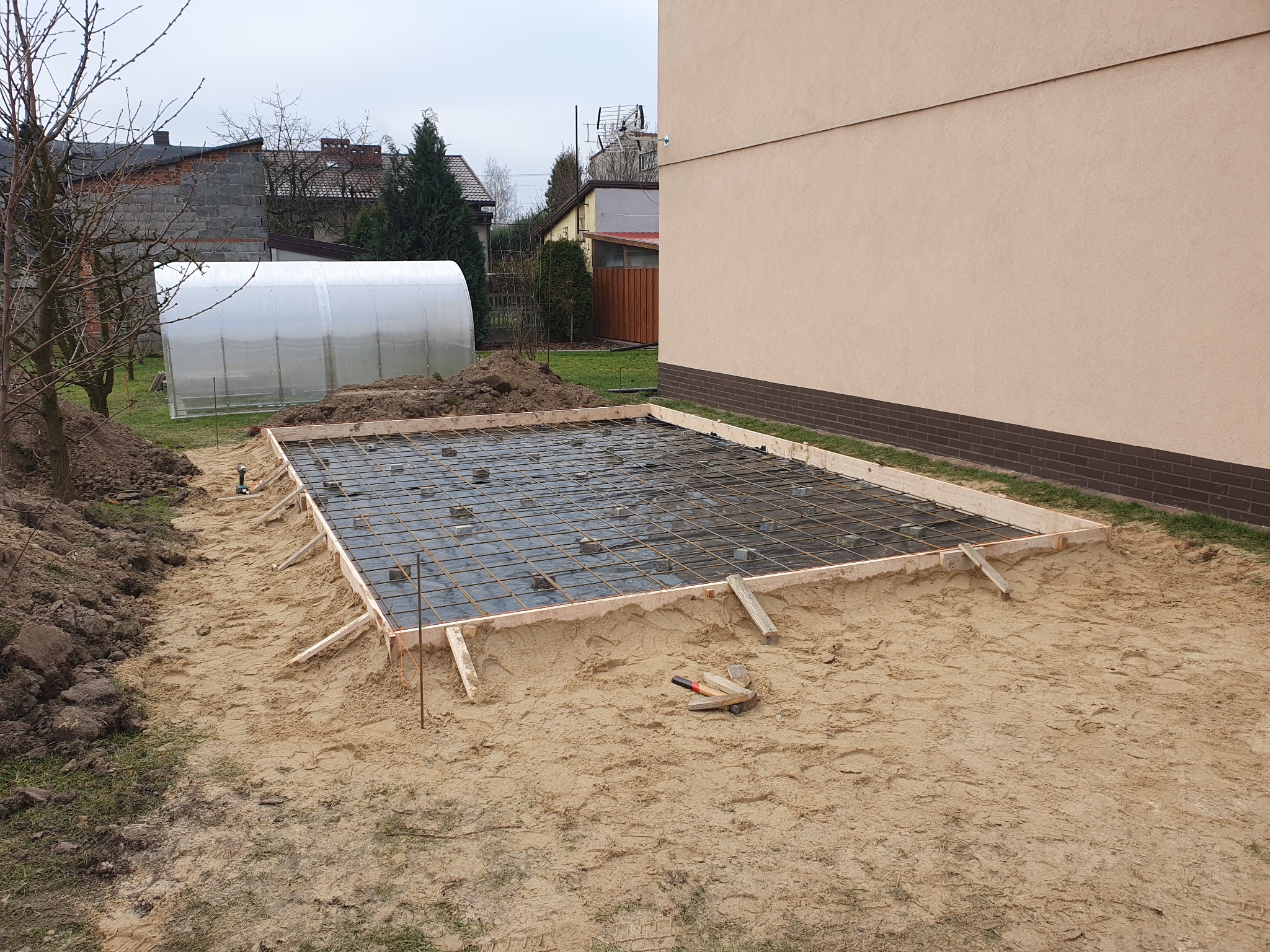 Przygotowany teren pod wylewkę fundamentu z deskowaniem, zbrojeniem i folią izolacyjną, widok z góry na piasek, w tle budynek mieszkalny i szklarnia.