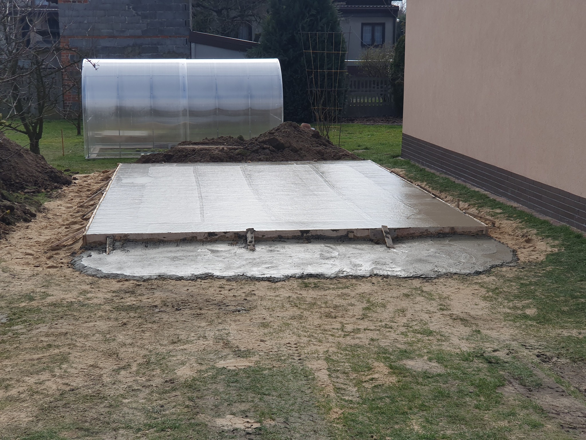 Świeżo wylana, mokra płyta betonowa na gruncie, otoczona drewnianym szalunkiem i ziemią, z widoczną folią pod spodem, w tle szklarnia i budynek.