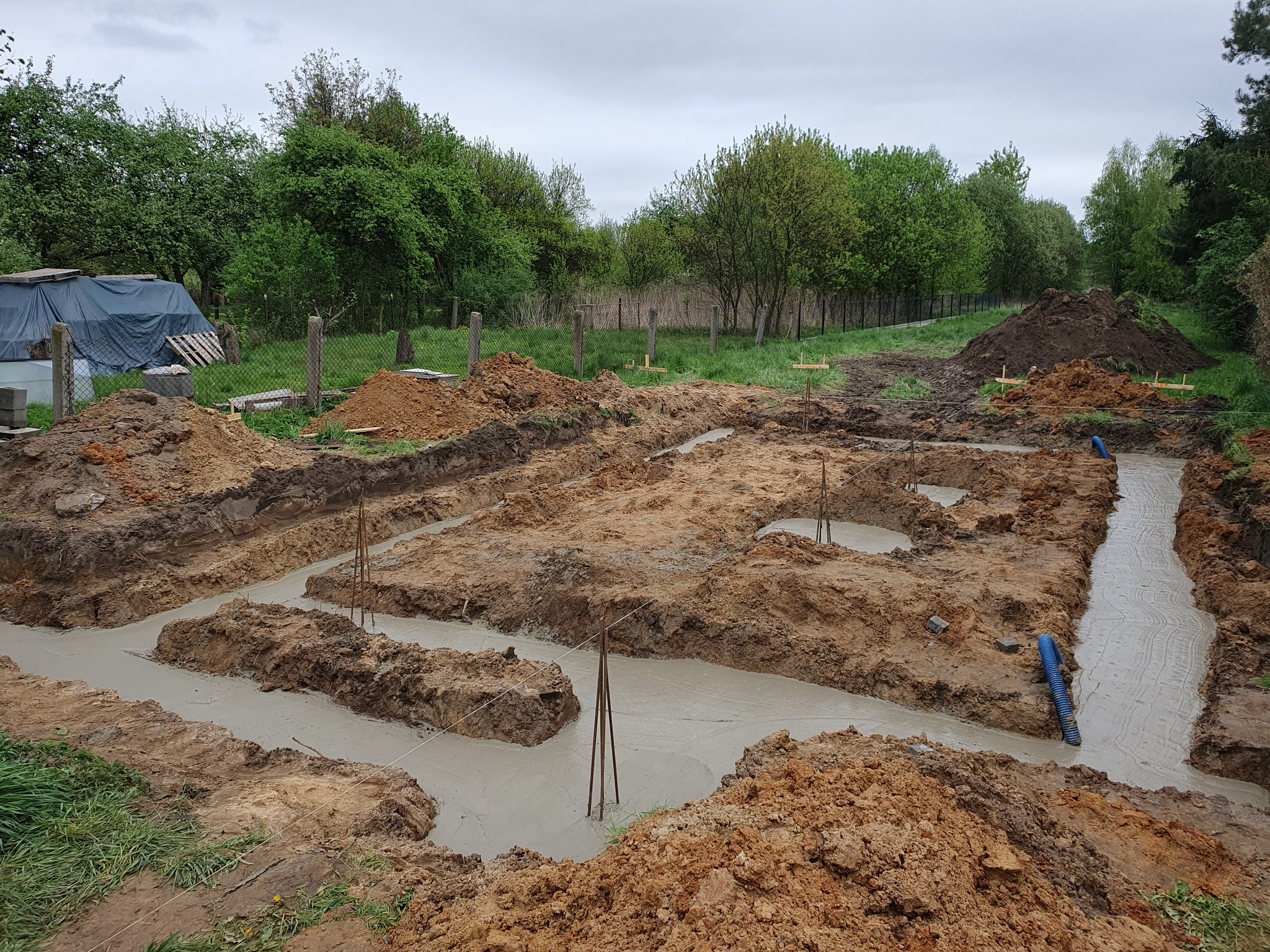 Wylane fundamenty z widocznymi prętami zbrojeniowymi i niebieską rurą drenażową, otoczone ziemią i zielenią w tle.