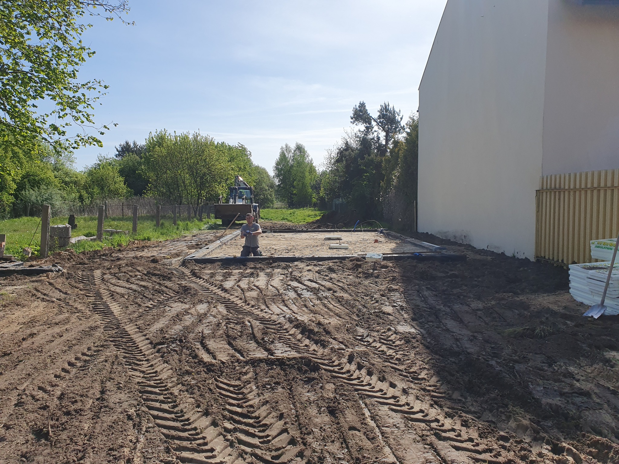 Przygotowanie terenu pod fundamenty: pracownik klęczy wewnątrz obrysu fundamentów, widoczne ślady opon koparki na ziemi, biały budynek w tle.
