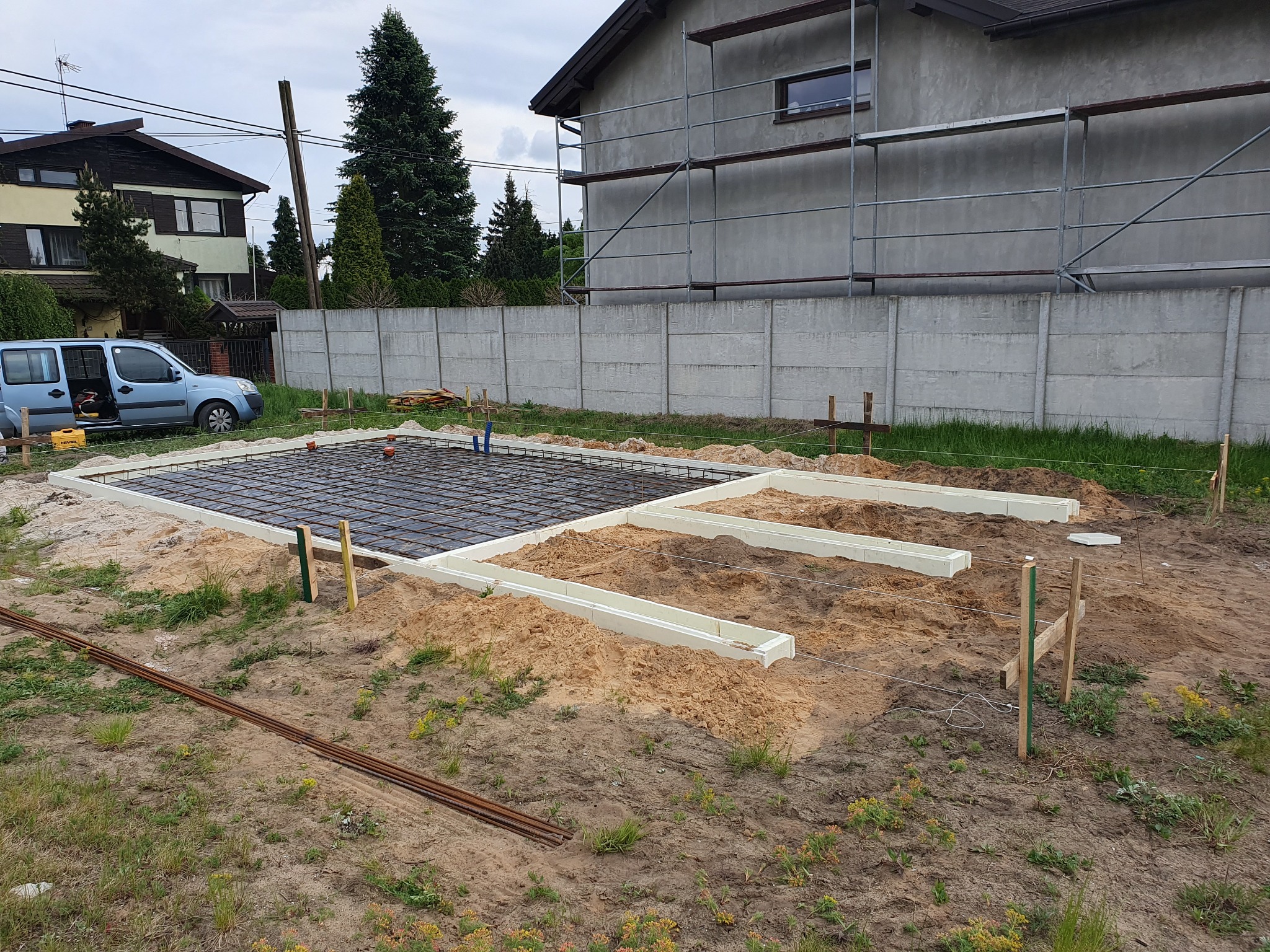 Przygotowany zbrojony szkielet pod wylewkę fundamentów na działce budowlanej, widoczne szalunki z białego styropianu, ułożone pręty zbrojeniowe oraz wystające pionowo rurki instalacyjne.