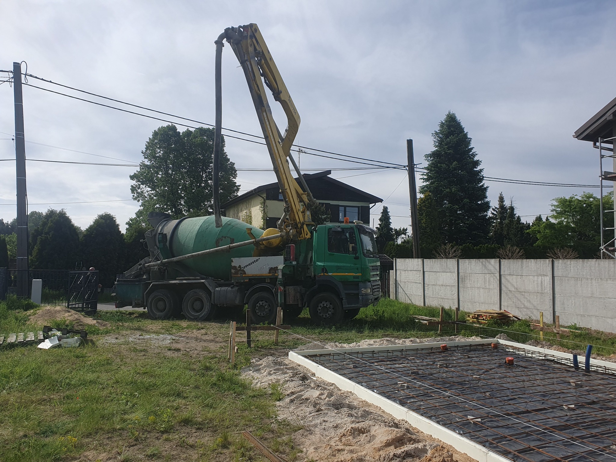 Betonomieszarka z wysięgnikiem pompuje beton na przygotowane zbrojenie fundamentu, widoczne szalunki i folia izolacyjna.