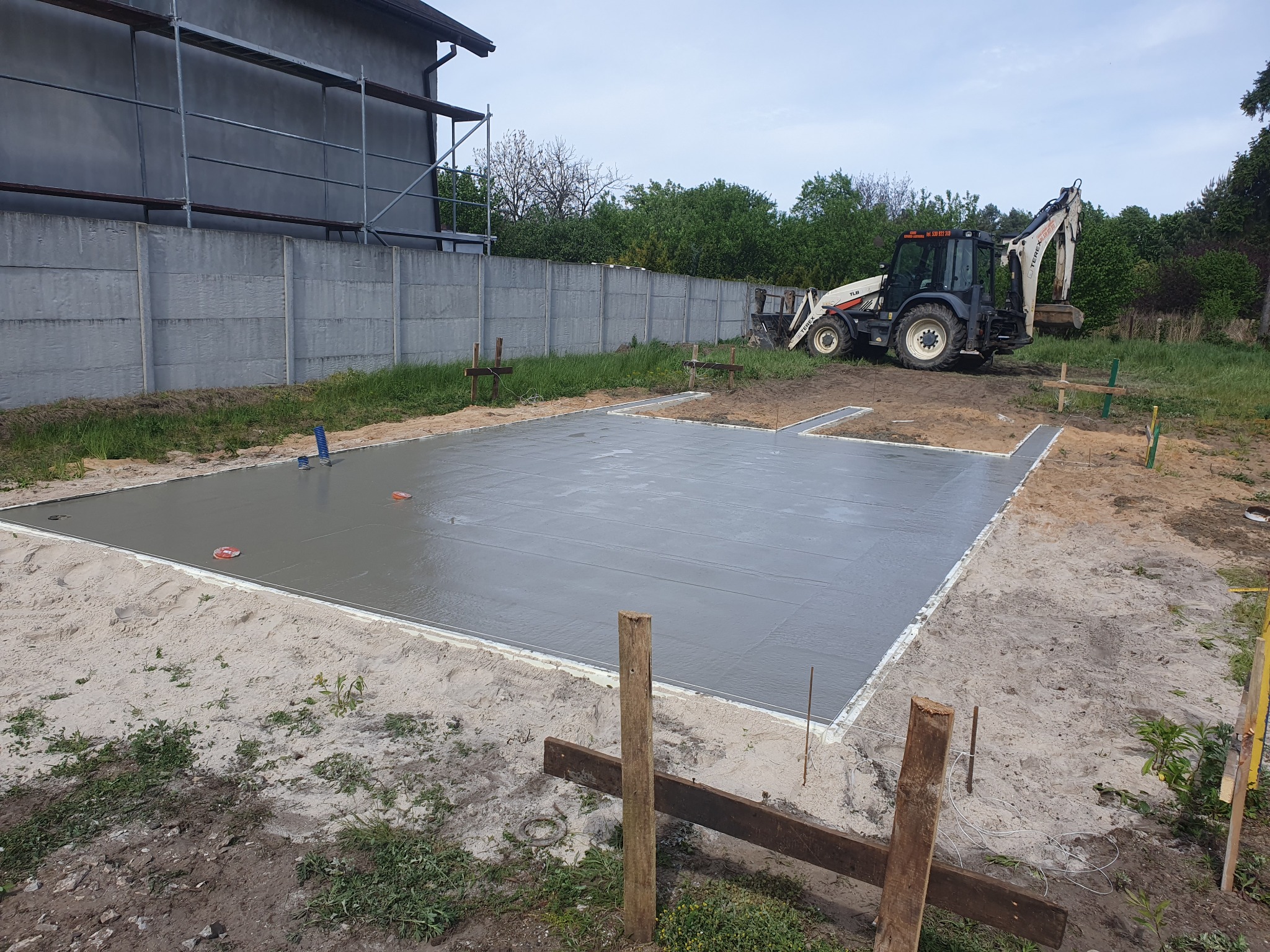 Świeżo wylana, gładka płyta fundamentowa na piaszczystym podłożu, obok stoi koparko-ładowarka, a w tle widać szary budynek w rusztowaniach i betonowy płot.