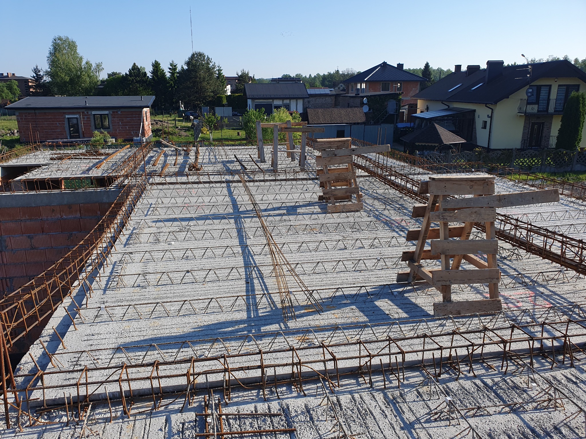 Zbrojenie stropu budynku jednorodzinnego z widocznymi podporami szalunkowymi, przygotowane do zalania betonem, w tle zabudowa mieszkalna.
