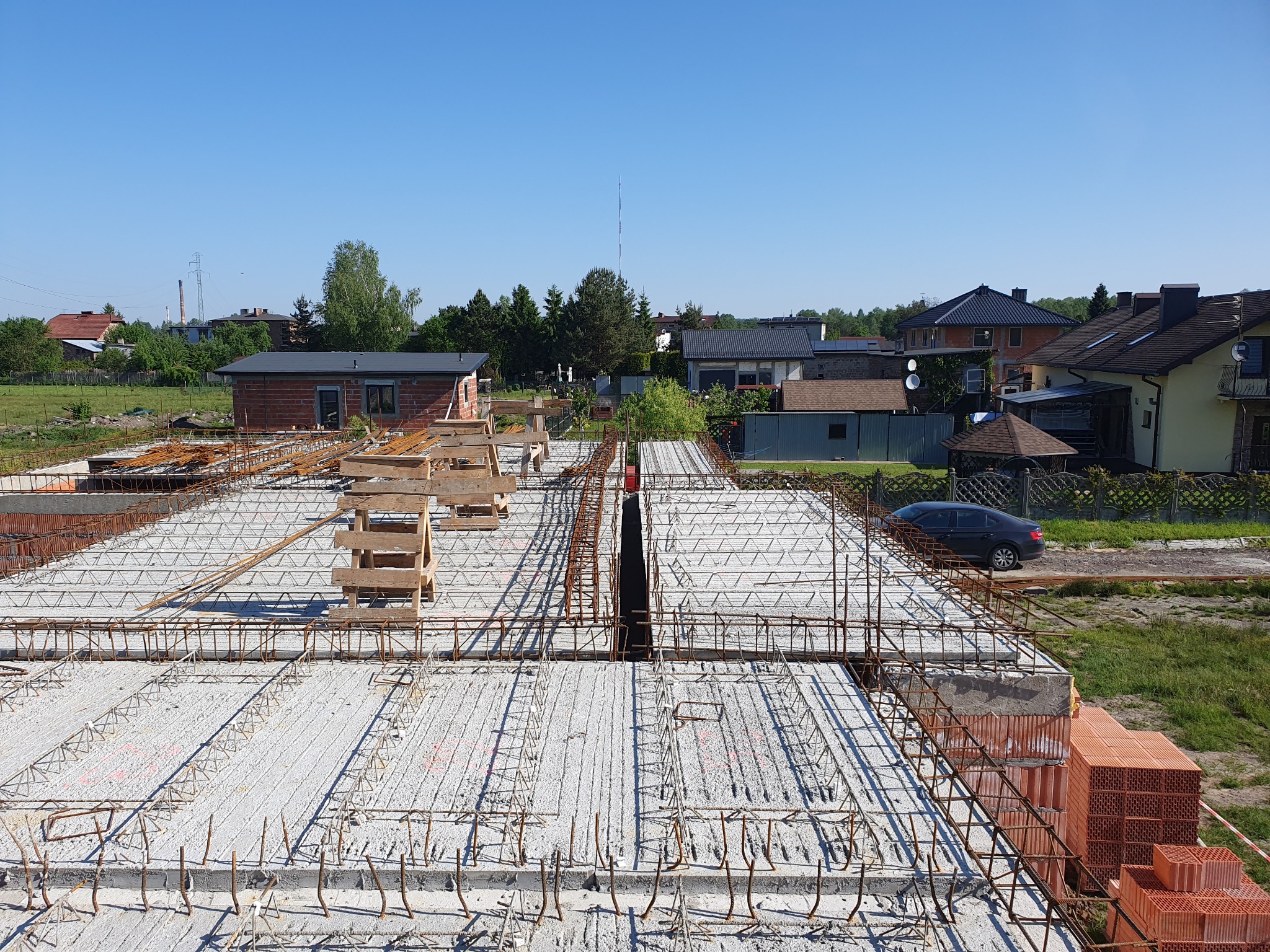 Zbrojenie stropu Teriva na budowie domu jednorodzinnego, widok z góry na ułożone pustaki betonowe i stalowe pręty, w tle zabudowa mieszkalna i błękitne niebo.