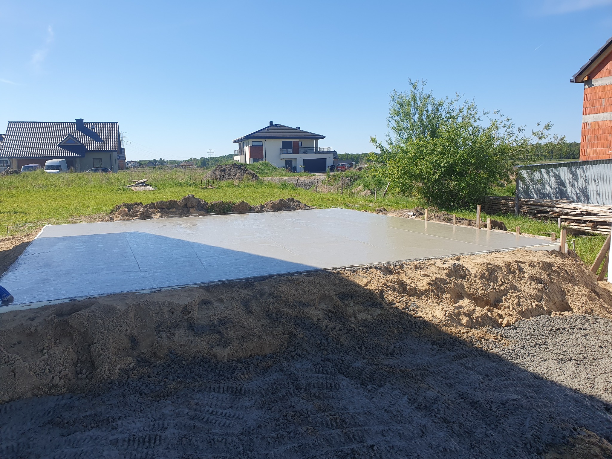 Świeżo wylana, gładka płyta fundamentowa z widocznym szalunkiem drewnianym, otoczona ziemią i gruzem, z domami w tle.