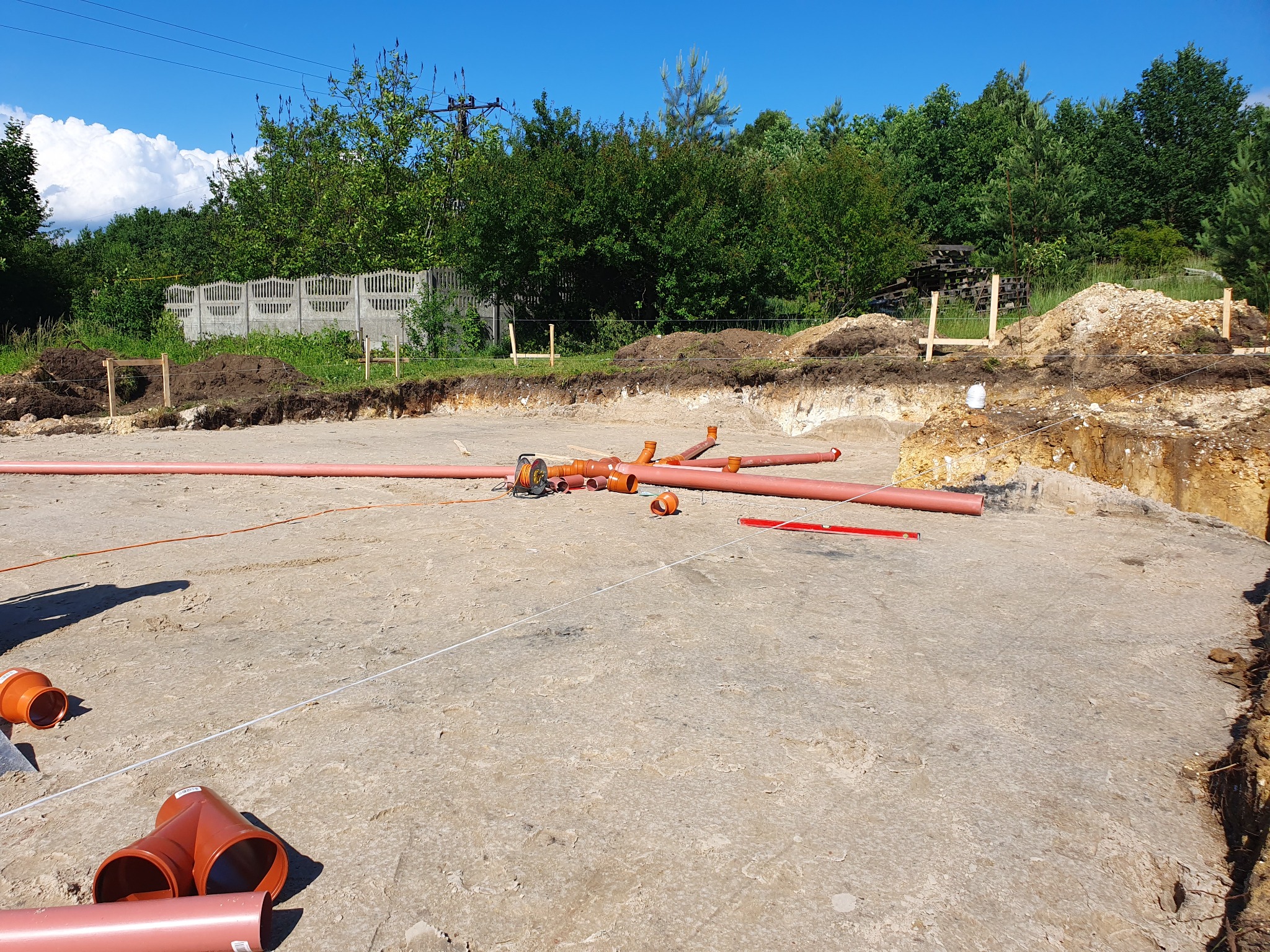 Przygotowanie terenu pod fundamenty w Postaszowicach: wykop z widocznymi rurami kanalizacyjnymi i narzędziami pomiarowymi na dnie wykopu, w tle ogrodzenie i zieleń.