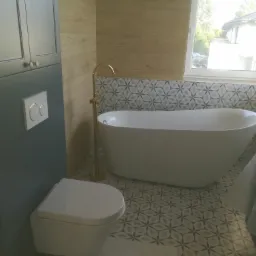 Nowoczesna łazienka z wolnostojącą wanną, złotą baterią podłogową, szarą szafką z umywalką podwieszaną i geometrycznymi płytkami podłogowymi oraz ściennymi.