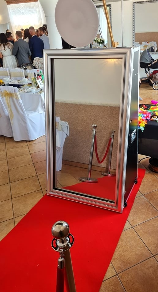 Lustrzana fotobudka z srebrną ramą ustawiona na czerwonym dywanie, widoczna w tle sala bankietowa z gośćmi.