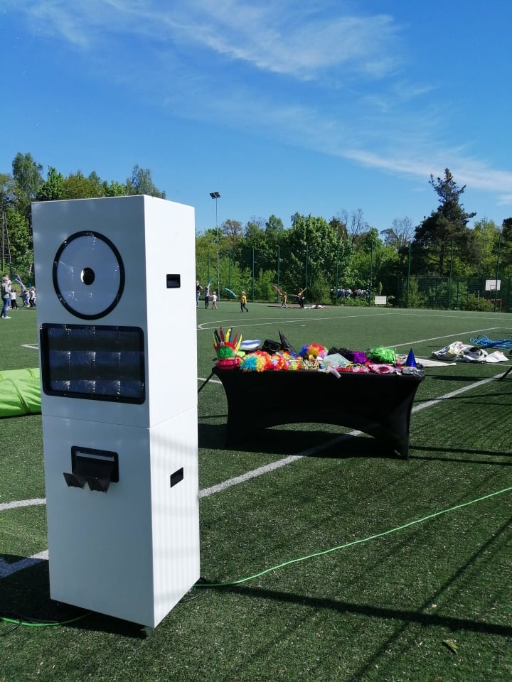 Biała fotobudka na tle zielonego boiska, obok stolik z kolorowymi rekwizytami, w tle drzewa i bawiące się dzieci.