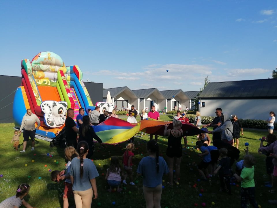 Dzieci bawią się na trawie, wokół kolorowe piłki, w tle dmuchany zamek ze zwierzętami i grupa dorosłych trzymających tęczową płachtę, błękitne niebo.