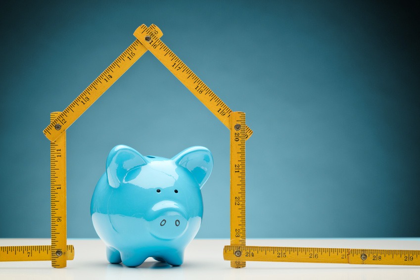 Niebieska świnka skarbonka pod konstrukcją z żółtej, składanej miarki w kształcie domu, na białym blacie i tle w odcieniach błękitu.