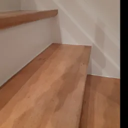 Fragment drewnianych schodów z białymi ścianami, widoczne niedoskonałości w wykończeniu spoin.