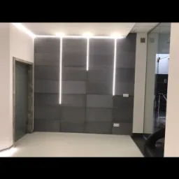 Nowoczesna ściana z szarych paneli z trzema pionowymi liniami świetlnymi LED, białe ściany boczne i sufit z listwą LED, widoczne gniazdka elektryczne i ościeżnica drzwiowa.