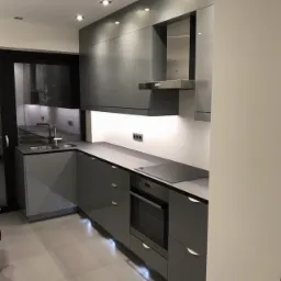 Nowoczesna kuchnia w odcieniach szarości z błyszczącymi frontami, oświetleniem podszafkowym i punktowym oraz płytkami podłogowymi w jasnym odcieniu szarości.